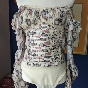 May include: Off-the-shoulder bodysuit with long sleeves. The top features a floral pattern in cream, pink, and black. The bodice has a smocked design, and the sleeves have a ruffled edge. The bottom is a solid cream color.