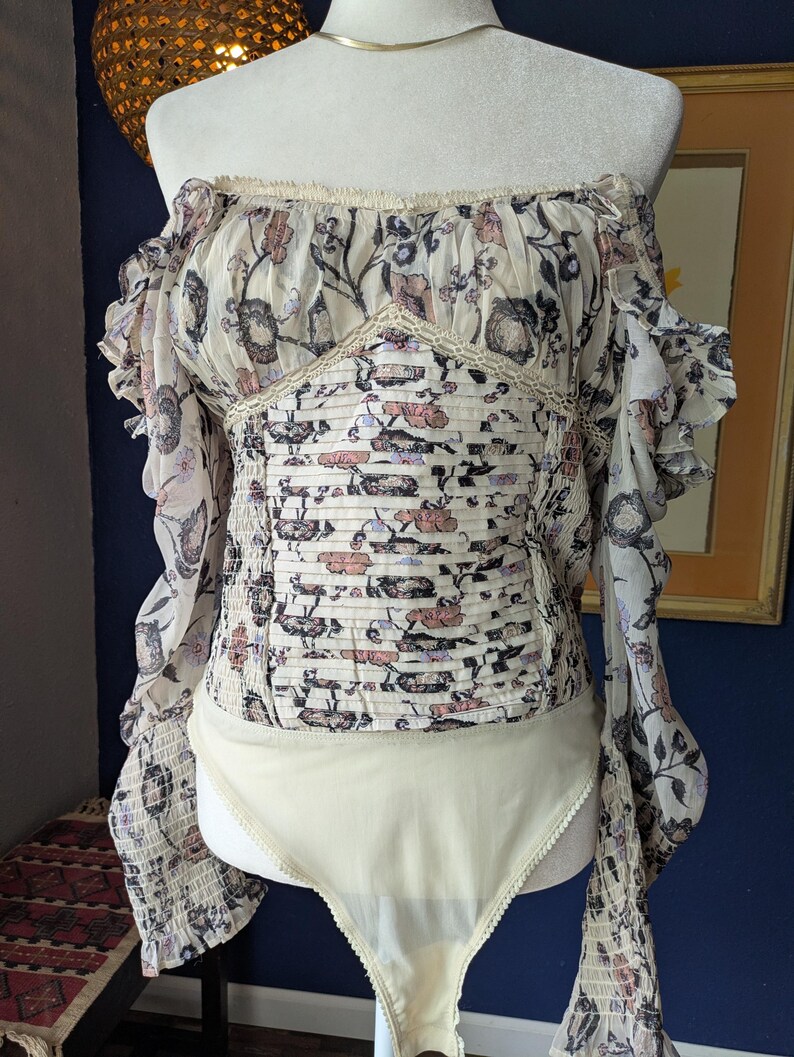 May include: Off-the-shoulder bodysuit with long sleeves. The top features a floral pattern in cream, pink, and black. The bodice has a smocked design, and the sleeves have a ruffled edge. The bottom is a solid cream color.