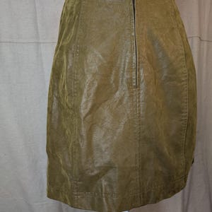 May include: Olive green leather skirt with a back zipper closure. The skirt has vertical seams and a slightly textured appearance. The garment is displayed on a white mannequin, showcasing its shape and design.