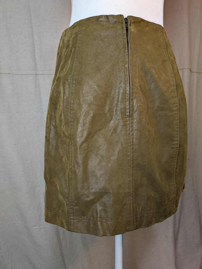 May include: Olive green leather skirt with a back zipper closure. The skirt has vertical seams and a slightly textured appearance. The garment is displayed on a white mannequin, showcasing its shape and design.