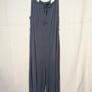 May include: A dark gray jumpsuit with a halter neckline and a drawstring detail. The sleeveless garment hangs on a wooden hanger against a neutral background. The jumpsuit features wide legs and a flowing silhouette.