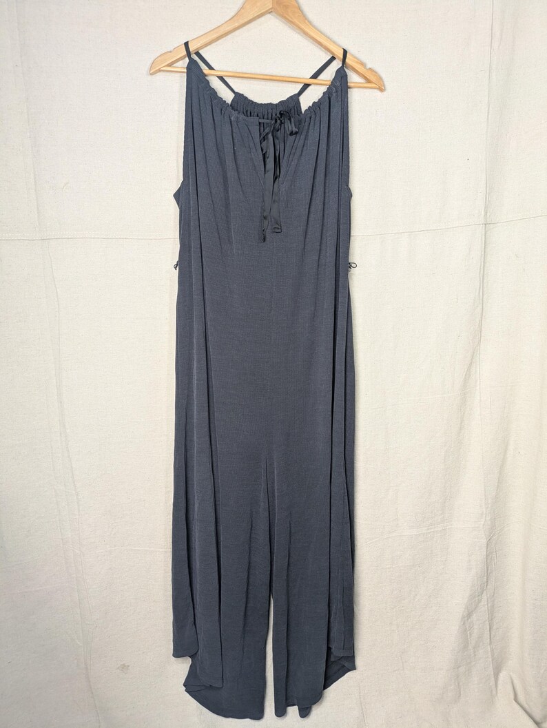 May include: A dark gray jumpsuit with a halter neckline and a drawstring detail. The sleeveless garment hangs on a wooden hanger against a neutral background. The jumpsuit features wide legs and a flowing silhouette.