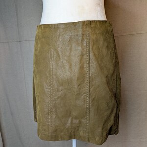 May include: Olive green mini skirt made of a textured material. The skirt has vertical seams down the front and back. The skirt is displayed on a white mannequin torso.