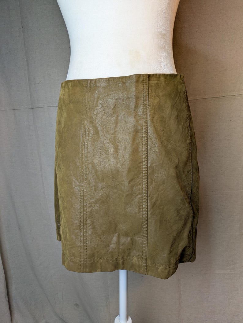 May include: Olive green mini skirt made of a textured material. The skirt has vertical seams down the front and back. The skirt is displayed on a white mannequin torso.