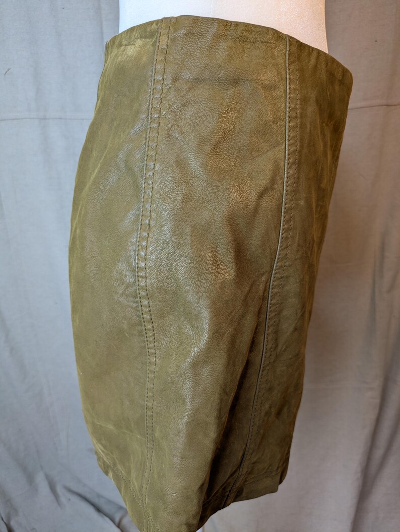 May include: A close-up of a khaki-colored leather skirt. The skirt has vertical seams down the front and back, with visible stitching. The leather has a slightly textured appearance.