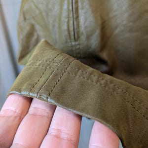 May include: Close-up of a section of an olive green leather jacket. The image highlights the stitching detail on the cuff and the texture of the leather. The jacket appears to be new, with a zipper visible in the background.