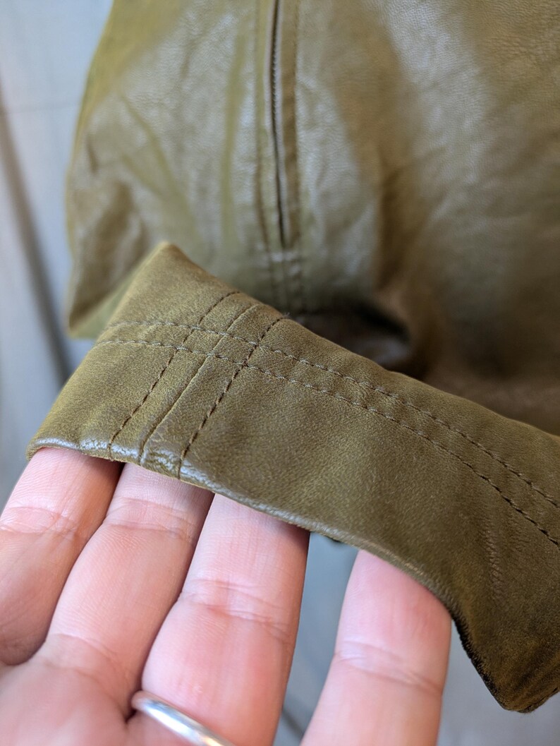 May include: Close-up of a section of an olive green leather jacket. The image highlights the stitching detail on the cuff and the texture of the leather. The jacket appears to be new, with a zipper visible in the background.