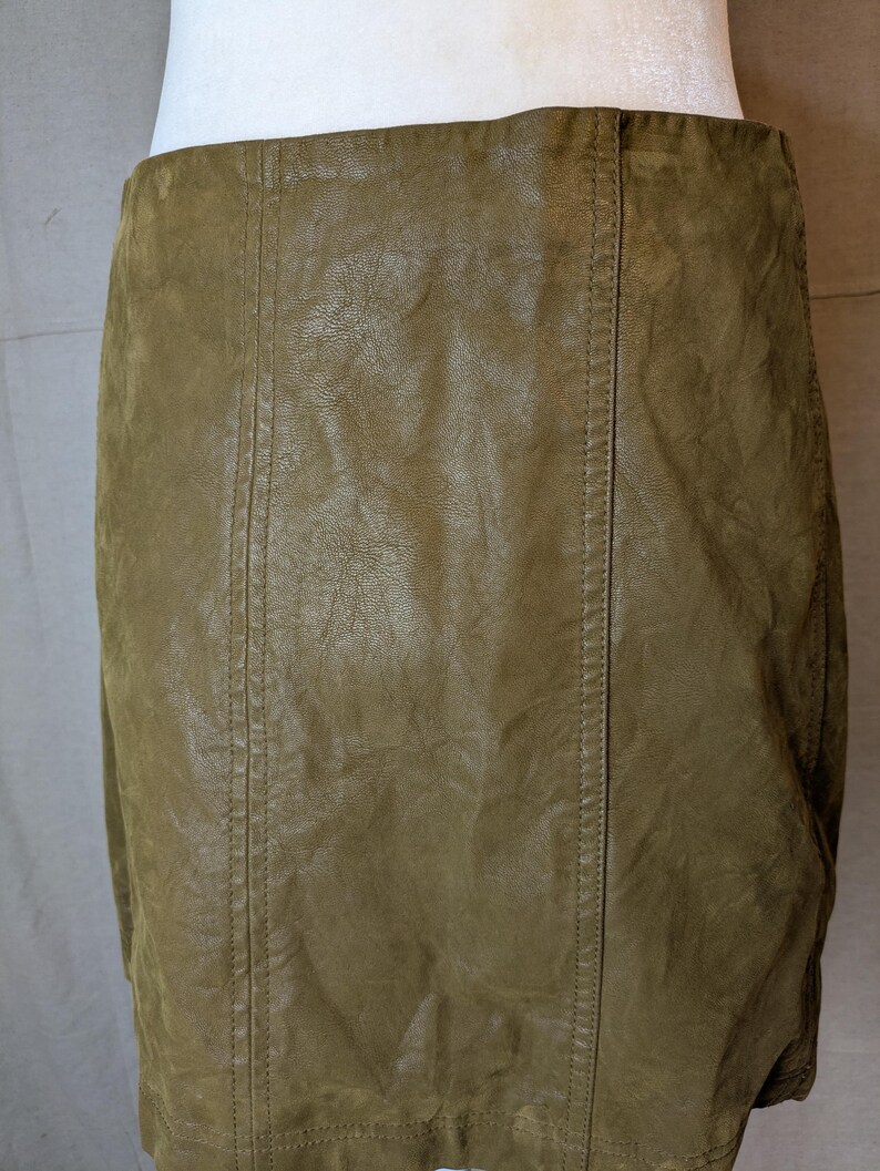 May include: Olive green leather skirt with vertical seam detailing. The skirt has a slightly textured appearance and a classic, fitted silhouette. The stitching is visible along the seams.