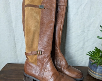 May include: A pair of knee-high brown leather boots with a suede panel. The boots have a buckle detail and a low heel. The boots are displayed on a wooden surface.