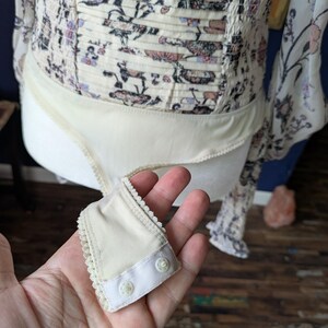 May include: Cream-colored bodysuit with a floral print bodice and long sleeves. The bodice has a smocked design. A small, cream-colored snap closure is held in the hand.