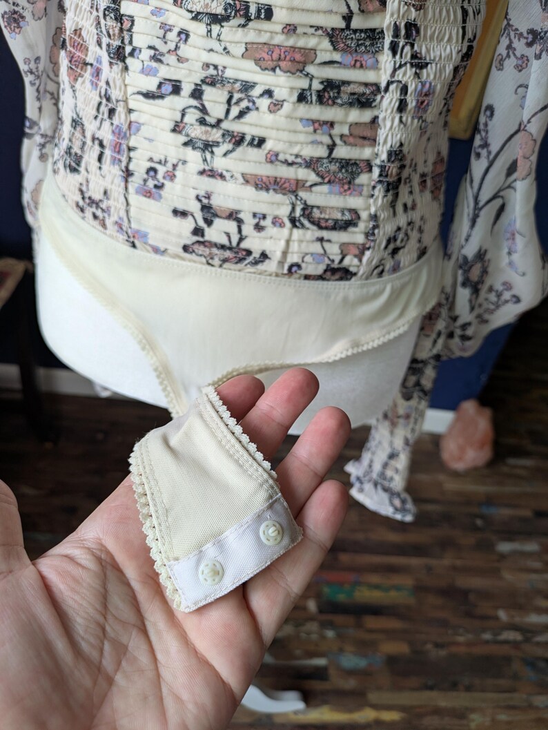 May include: Cream-colored bodysuit with a floral print bodice and long sleeves. The bodice has a smocked design. A small, cream-colored snap closure is held in the hand.