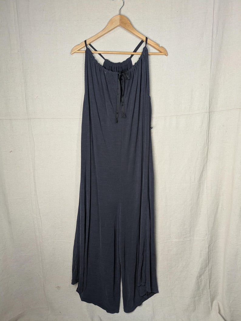 May include: A dark gray jumpsuit with a halter neckline and wide legs hangs from a wooden hanger. The fabric appears soft and flowing, with a drawstring detail at the neckline. The jumpsuit is a solid color, suitable for casual or semi-formal wear.