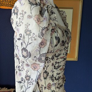 May include: A cream-colored blouse with a floral pattern in shades of pink, gray, and black. The blouse features long, sheer sleeves and a fitted bodice with a smocked design. The neckline is ruffled.