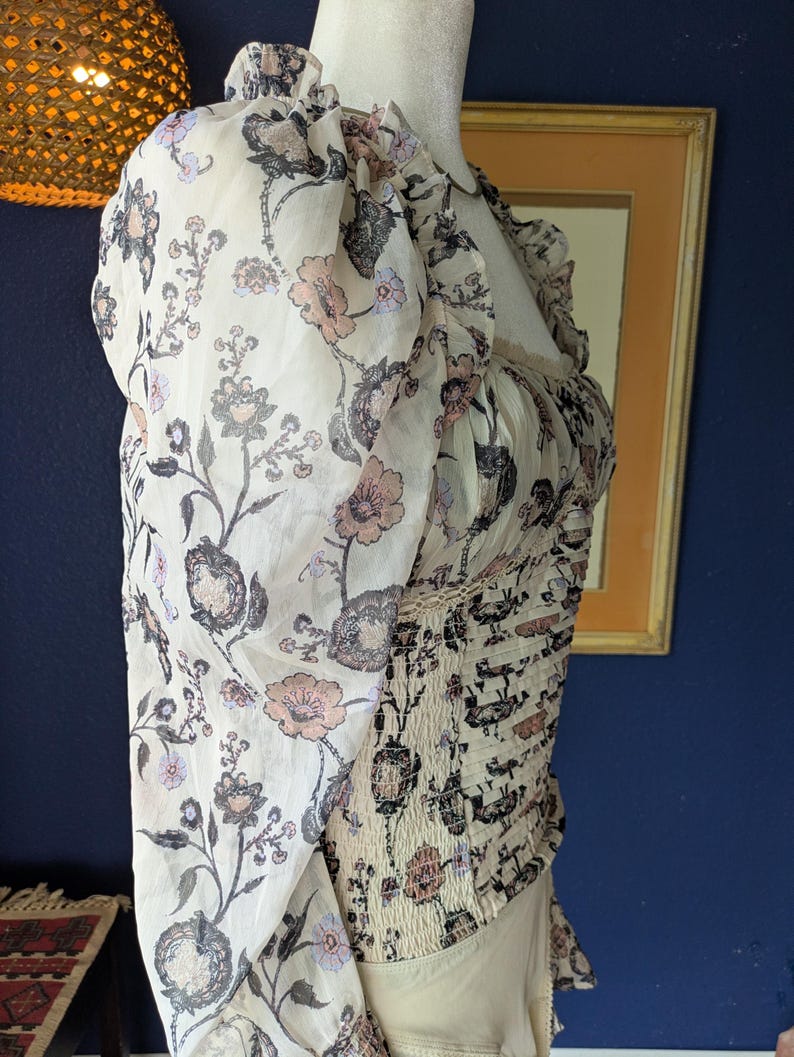 May include: A cream-colored blouse with a floral pattern in shades of pink, gray, and black. The blouse features long, sheer sleeves and a fitted bodice with a smocked design. The neckline is ruffled.