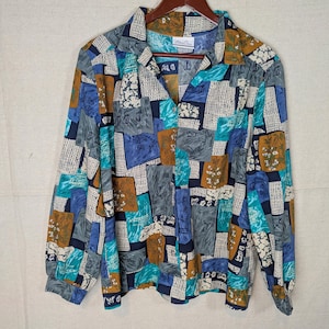 May include: A long-sleeved button-down shirt with a colorful patchwork design. The shirt features a V-neck collar and a pattern of squares in shades of blue, teal, brown, and gray. The shirt is hanging on a wooden hanger.