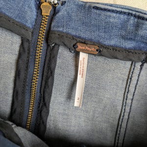 May include: Close-up of a blue denim garment with a brass zipper and a "Free People" tag. The tag indicates the garment was made in Egypt and is a size 2. The zipper is centered, and the denim has visible stitching.