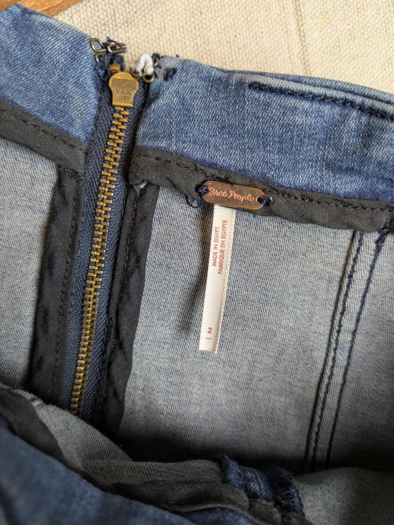 May include: Close-up of a blue denim garment with a brass zipper and a "Free People" tag. The tag indicates the garment was made in Egypt and is a size 2. The zipper is centered, and the denim has visible stitching.