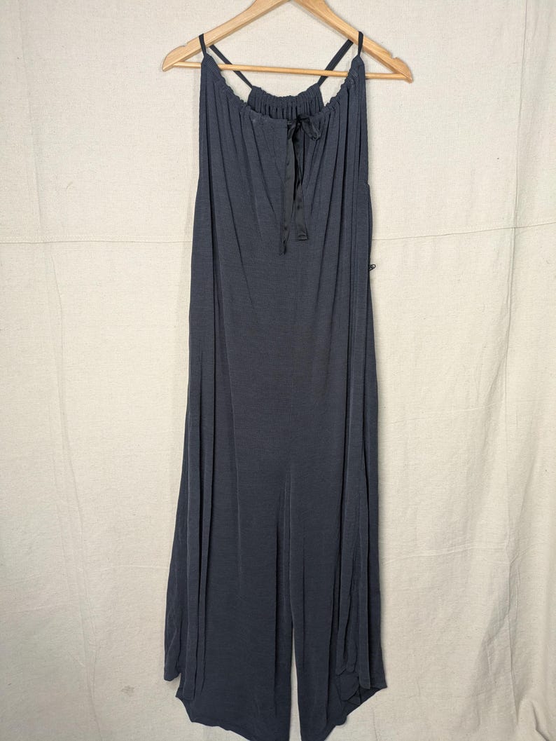 May include: A dark gray jumpsuit with a halter neckline and a drawstring detail. The jumpsuit has wide legs and is hanging on a wooden hanger. The fabric appears to be soft and flowing.
