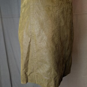 May include: Olive green faux leather skirt. The skirt is a mini-skirt style with vertical seams down the front and back. The skirt is displayed on a white mannequin.