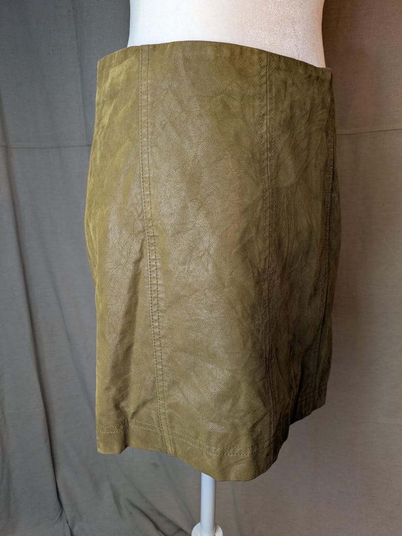 May include: Olive green faux leather skirt. The skirt is a mini-skirt style with vertical seams down the front and back. The skirt is displayed on a white mannequin.