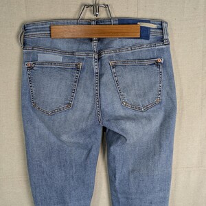 May include: Light blue denim jeans with a classic five-pocket design, visible stitching, and a faded wash. The jeans are hanging on a wooden hanger, showcasing the back pockets and waistband details. The denim appears to be a medium weight.