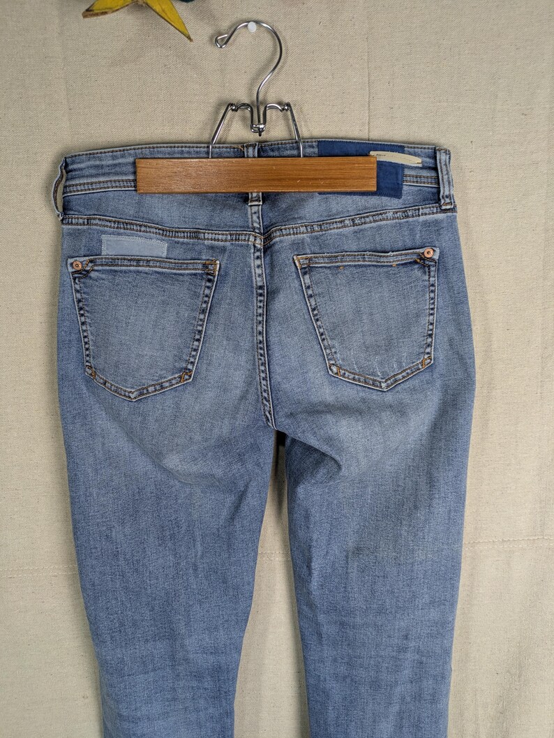 May include: Light blue denim jeans with a classic five-pocket design, visible stitching, and a faded wash. The jeans are hanging on a wooden hanger, showcasing the back pockets and waistband details. The denim appears to be a medium weight.
