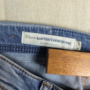 May include: Close-up of blue denim jeans with a light wash. The jeans have visible stitching in a contrasting color and a label that reads "Pilcro" and "by Anthropologie". A wooden hanger is partially visible.