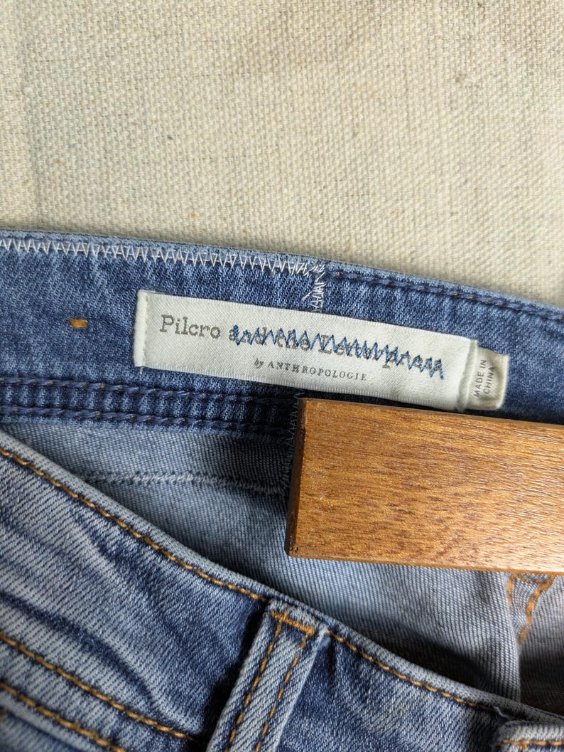 May include: Close-up of blue denim jeans with a light wash. The jeans have visible stitching in a contrasting color and a label that reads "Pilcro" and "by Anthropologie". A wooden hanger is partially visible.