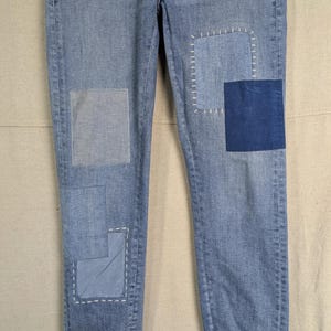 May include: Light blue denim jeans with multiple patches of varying shades of blue. Patches are sewn on with white thread. The cuffs are rolled up, revealing a brown and orange trim. The jeans are a classic, casual style.