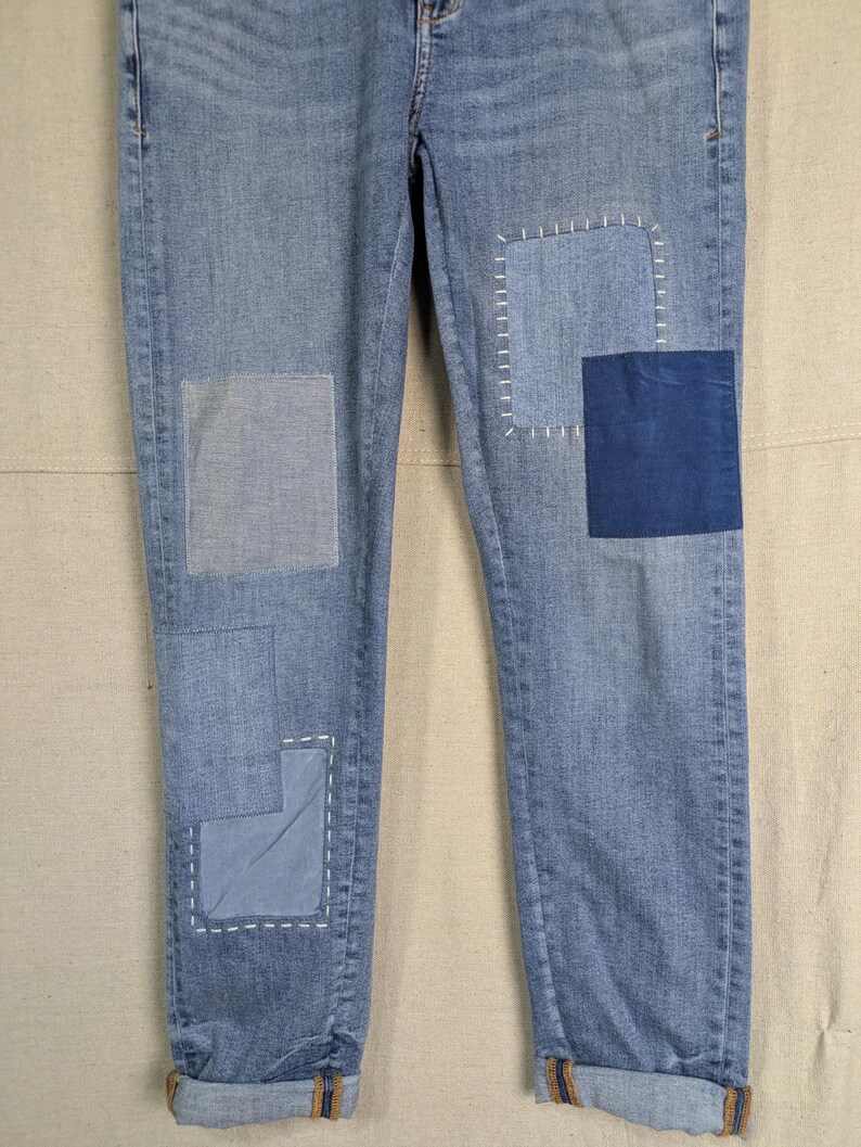 May include: Light blue denim jeans with multiple patches of varying shades of blue. Patches are sewn on with white thread. The cuffs are rolled up, revealing a brown and orange trim. The jeans are a classic, casual style.