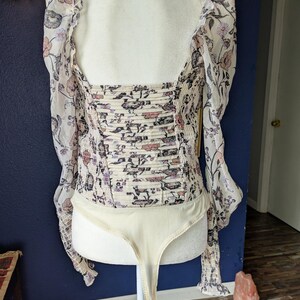 May include: A cream-colored bodysuit with a floral pattern in pink, purple, and black. The long sleeves are sheer, and the bodice has a ruched design. The bodysuit is displayed on a mannequin.
