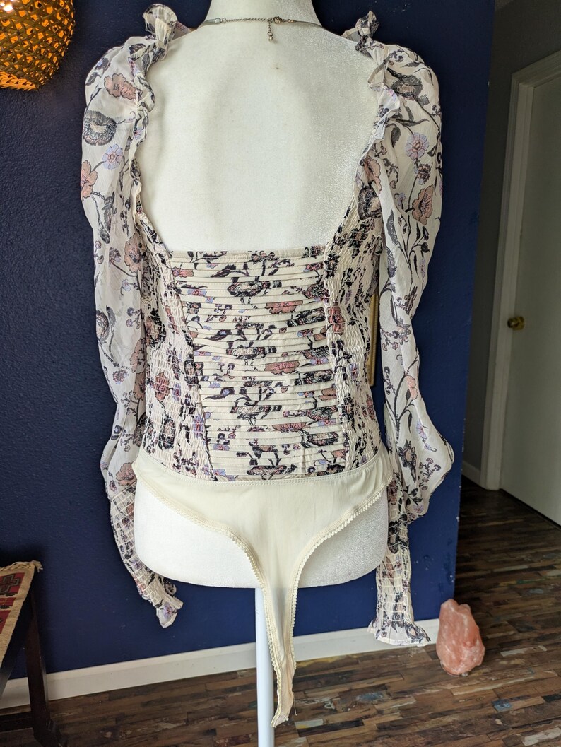 May include: A cream-colored bodysuit with a floral pattern in pink, purple, and black. The long sleeves are sheer, and the bodice has a ruched design. The bodysuit is displayed on a mannequin.