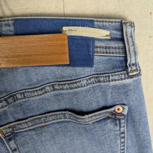 May include: Close-up of light blue denim jeans with a blue patch and a wooden hanger. The jeans have a back pocket with a copper-colored rivet. The tag reads "Pilcro and the Letterpress."