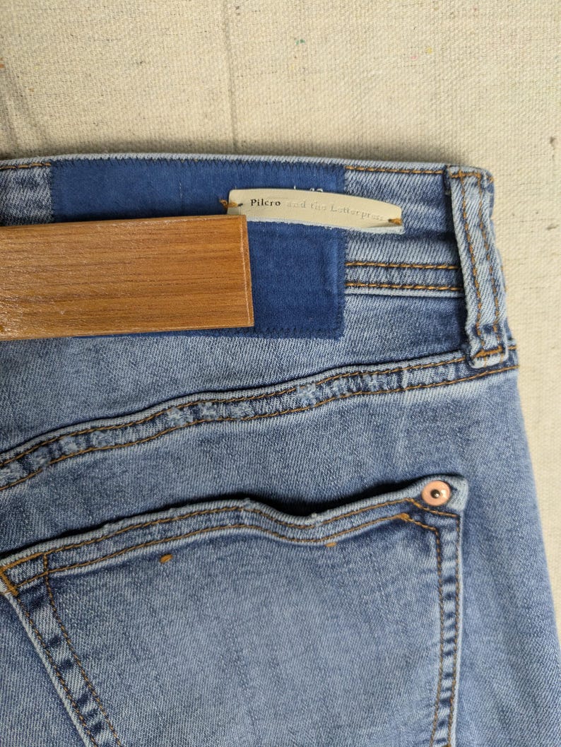 May include: Close-up of light blue denim jeans with a blue patch and a wooden hanger. The jeans have a back pocket with a copper-colored rivet. The tag reads "Pilcro and the Letterpress."