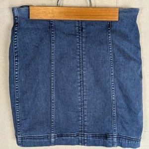 May include: A denim skirt in a medium blue wash, hanging on a wooden hanger. The skirt features vertical seams and a classic design. The garment is made of a sturdy denim material.