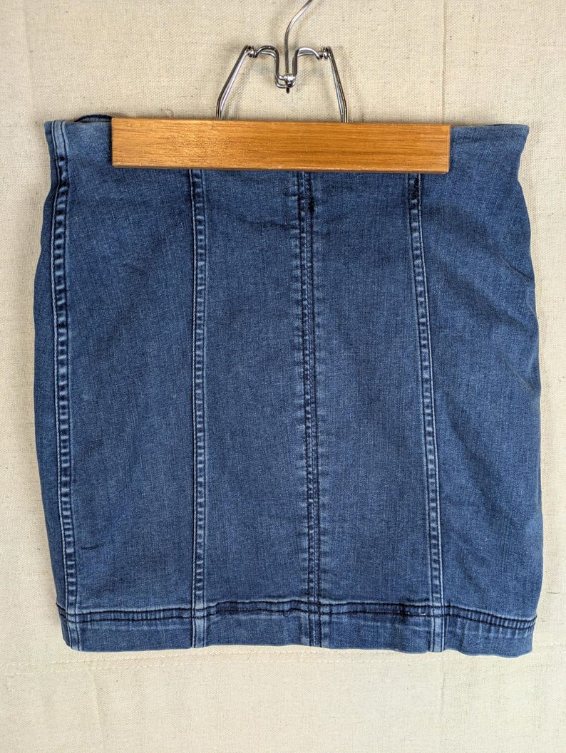 May include: A denim skirt in a medium blue wash, hanging on a wooden hanger. The skirt features vertical seams and a classic design. The garment is made of a sturdy denim material.