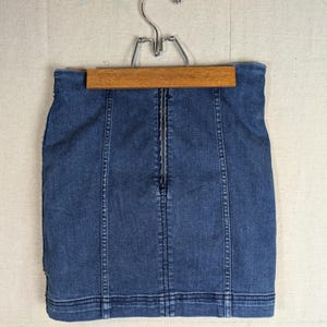 May include: A denim mini skirt in a medium blue wash, hanging on a wooden hanger. The skirt features a front zipper closure and vertical seam detailing. The garment is a classic style, suitable for casual wear.