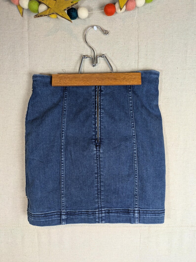 May include: A denim mini skirt in a medium blue wash, hanging on a wooden hanger. The skirt features a front zipper closure and vertical seam detailing. The garment is a classic style, suitable for casual wear.