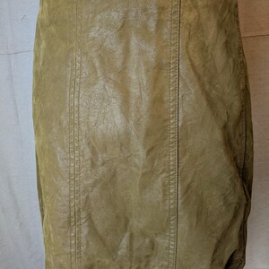 May include: A tan-colored, faux leather skirt. The skirt has a straight cut and features vertical seams down the front and back. The material has a textured appearance, and the skirt appears to be worn.