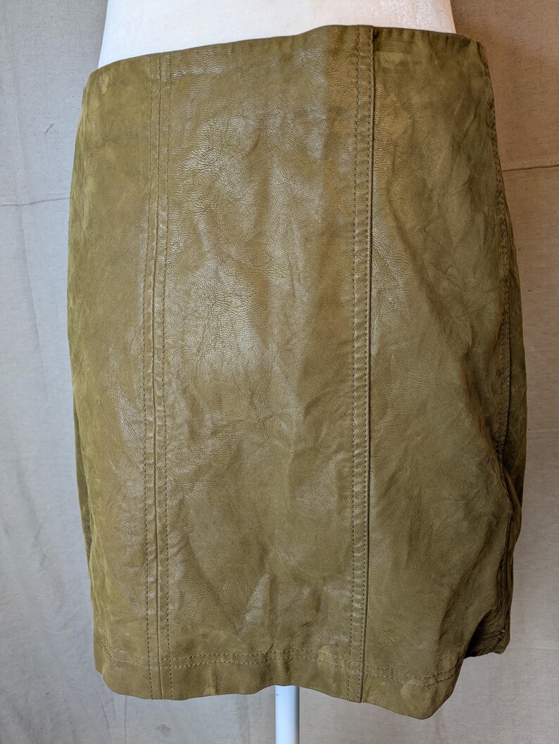 May include: A tan-colored, faux leather skirt. The skirt has a straight cut and features vertical seams down the front and back. The material has a textured appearance, and the skirt appears to be worn.