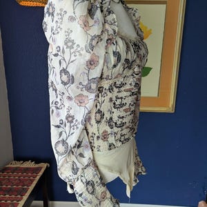 May include: A cream-colored bodysuit with a floral pattern in shades of pink, gray, and black. The top features sheer, puffed sleeves and a ruffled neckline. The bodice has a fitted, smocked design.