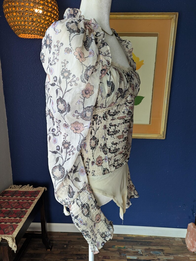 May include: A cream-colored bodysuit with a floral pattern in shades of pink, gray, and black. The top features sheer, puffed sleeves and a ruffled neckline. The bodice has a fitted, smocked design.