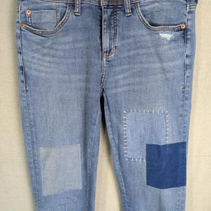 May include: Light blue denim jeans with patches of varying shades of blue. The jeans have a classic five-pocket design and are hanging on a wooden hanger. The patches are sewn on with white thread, adding a unique, handcrafted look.