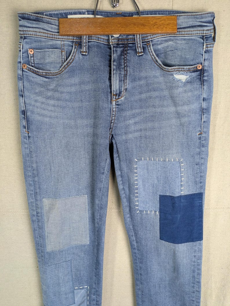 May include: Light blue denim jeans with patches of varying shades of blue. The jeans have a classic five-pocket design and are hanging on a wooden hanger. The patches are sewn on with white thread, adding a unique, handcrafted look.