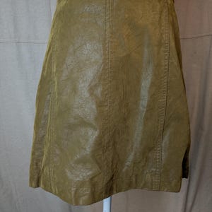 May include: Olive green faux leather skirt. The skirt has a slight A-line shape and features vertical seams down the front and back. The skirt is displayed on a white mannequin.