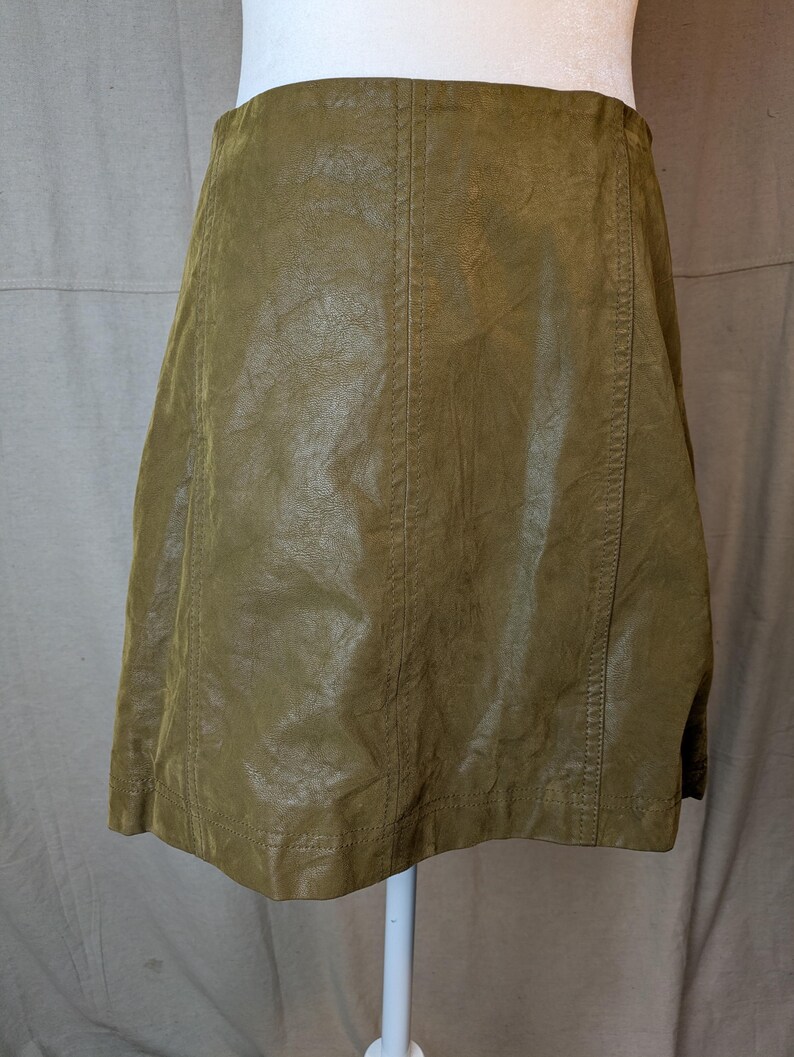 May include: Olive green faux leather skirt. The skirt has a slight A-line shape and features vertical seams down the front and back. The skirt is displayed on a white mannequin.