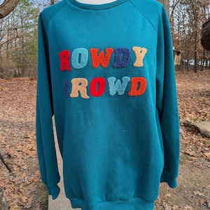 May include: Teal sweatshirt with the word "ROWDY ROWD" in colorful, textured letters across the chest. The sweatshirt has a crew neck and long sleeves. The letters are in shades of red, orange, blue, and yellow.