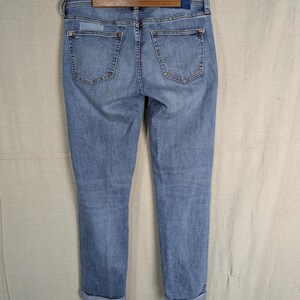 May include: Light blue denim jeans with a classic five-pocket design. The jeans are cuffed at the ankles and hang from a wooden hanger. The back pockets and waistband are visible, showcasing the stitching and details.