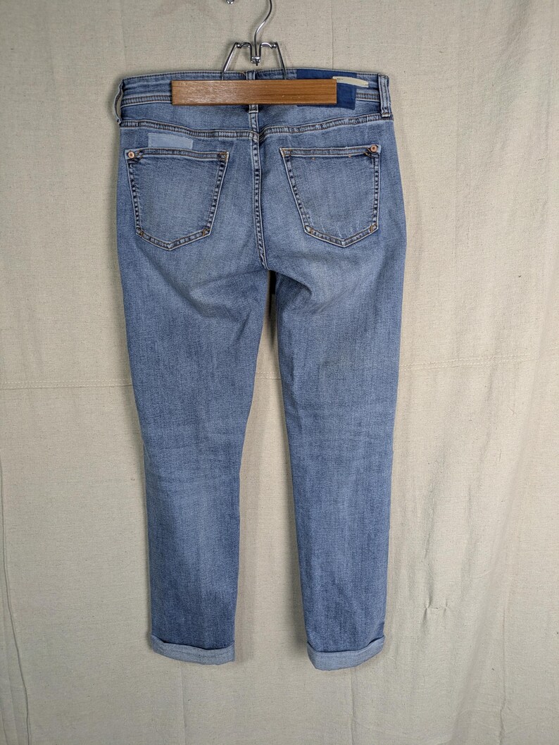 May include: Light blue denim jeans with a classic five-pocket design. The jeans are cuffed at the ankles and hang from a wooden hanger. The back pockets and waistband are visible, showcasing the stitching and details.