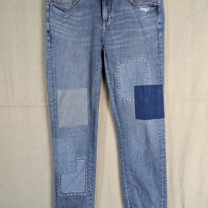 May include: Light blue denim jeans with patches of varying shades of blue. The jeans have a distressed look with a small rip on the right knee. The cuffs are rolled up, revealing a contrasting fabric.